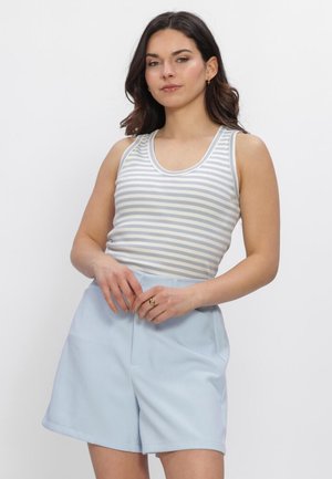 Jonge vrouw met donker haar draagt een mouwloze witte en grijze gestreepte top en lichtblauwe shorts met hoge taille, staand met handen gevouwen.