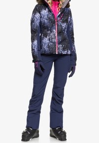 Puffige Winterjacke mit einem dunklen, gesprenkelten Design, rosa Akzenten und einer mit Fell gefütterten Kapuze, kombiniert mit marineblauen Schneehosen und schwarzen Skischuhen.