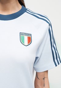 Chemise blanche avec col bleu et rayures, écusson de bouclier italien sur la poitrine, tatouage visible sur l'avant-bras de la personne.