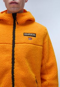 Ljusorange fleecejacka med texturerad yta, utrustad med en svart dragkedja och en liten patchlogotyp på bröstet. Med huva.