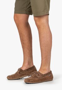 Zapatos de barco de ante marrón con cordones y ojales plateados, que presentan un upper texturizado y una suela de goma visible debajo de los pantalones cortos de longitud hasta el tobillo.