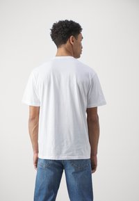 Tricou alb din bumbac, cu mâneci scurte, decolteu rotund, vedere din spate arătând o textură netedă și un croi lejer, asortat cu blugi din denim albastru.