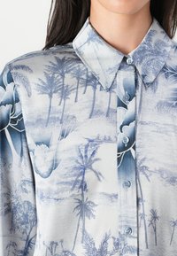 Camicia azzurra chiaro con bottoni, decorata con palme e disegni floreali. Il tessuto è liscio con una leggera lucentezza e presenta un colletto classico.