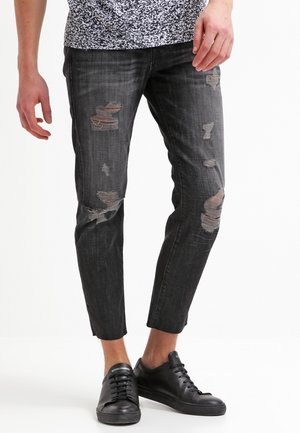 Osoba nosząca czarne, wąskie dżinsy z przetarciami i dziurami oraz czarne niskie sneakersy, stojąca na białym tle.