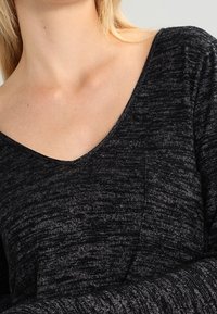 Top en V noir et gris foncé chiné, avec des manches longues et un détail de poche, ayant une apparence douce et texturée.