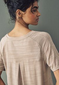 Top de deporte de color beige claro con tejido texturizado, mangas cortas, un corte relajado y un pliegue en la espalda; presenta el logo "ACTIVE" cerca del cuello.