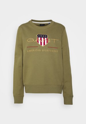 Olive green sweatshirt with crew neck, ribbed cuffs, and hem. Features embroidered shield logo with red and white stripes, "GANT" text, and "AMERICAN SPORTSWEAR".