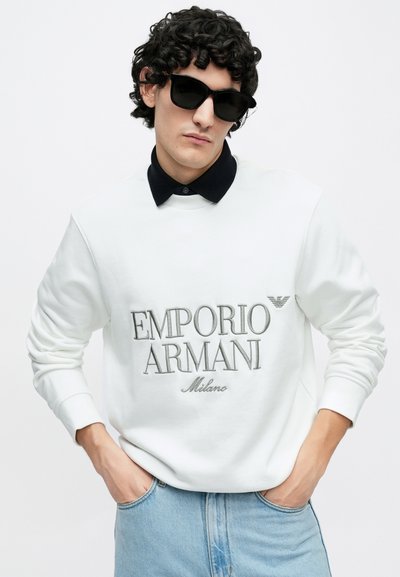 Sweatshirt branca com texto bordado "Emporio Armani Milano", combinada com uma camisa preta com gola por baixo. Usada com óculos de sol pretos.