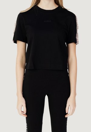 Kobieta w czarnym, krótkim T-shircie z różowymi paskami po bokach oraz czarnych legginsach z białym, firmowym napisem na lewej nodze.