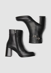 Bottines en cuir noir avec fermeture éclair sur le côté, talon carré et accents décoratifs en boucle. Texture lisse et design épuré.