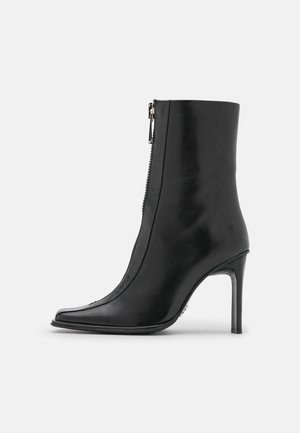 Bota de cuero negro hasta el tobillo con punta afilada, cremallera lateral y tacón alto y delgado, mostrada de perfil sobre un fondo liso.
