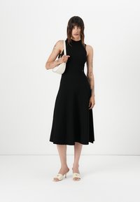 Svart ermeløs kjole med tettsittende bodice og utsvingt skjørt. Jevnt stoff, rund hals. Kombinert med en hvit veske og lyse sandaler.