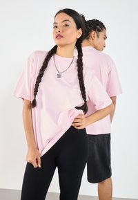 Lys rosa t-skjorte med korte ermer i løs passform, sammen med svarte ribbede leggings. En sølvkjede med anheng er brukt.