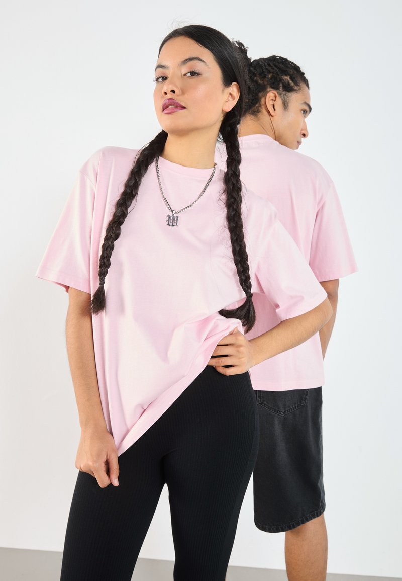Lys rosa t-skjorte med korte ermer i løs passform, sammen med svarte ribbede leggings. En sølvkjede med anheng er brukt.