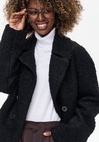 Czarny teksturowany płaszcz z dużymi klapami i dwoma guzikami, noszony na białym golfie z subtelnym wzorem, zestawiony z brązowymi spodniami z wysokim stanem.