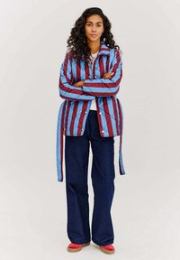 Překládaná bunda v červených a modrých pruzích se stojáčkem, bočními tkanicemi a patentkami, doplněná tmavými denimovými kalhotami a červenými botami.