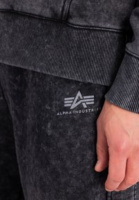Gros plan sur la main droite d'une personne reposant à côté d'un pantalon de survêtement Alpha Industries gris foncé et d'un sweat-shirt à col côtelé assorti.