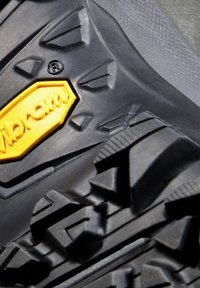Sort gummi yttersåle med dype, multirettede mønstre. Har en gul Vibram-logo på en teksturert seksjon. Holdbart design for godt grep.