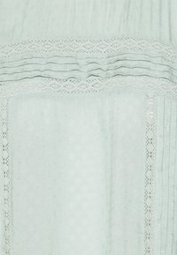 Helles mintgrünes Gewebe mit strukturierten Mustern, Spitzelementen und horizontalen Falten, das ein weiches, luftiges Aussehen und zarte Details aufweist.
