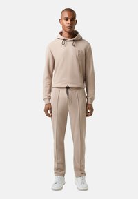 Beige hoodie met trekkoorden en logo patch, gecombineerd met bijpassende taps toelopende sweatpants met plooien. Gedragen met witte sneakers.