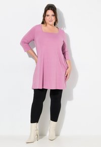 Rosa Tunika mit quadratischem Ausschnitt und drei Viertelärmeln, kombiniert mit schwarzen Leggings und hellbeigen, hohen Stiefeln. Glatte Stofftextur.