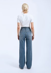Jeans de mezclilla azul de tiro alto y pierna ancha con dos bolsillos traseros y costuras visibles. Combinados con una camiseta blanca sencilla.