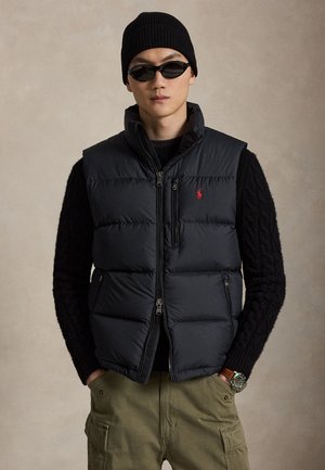 Uomo che indossa un berretto di maglia nero, occhiali da sole neri, giubbotto imbottito nero con logo rosso, maglione nero, pantaloni cargo oliva e orologio da polso con quadrante verde.