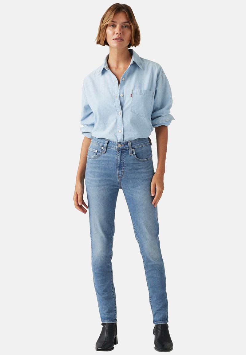 Jeans de mezclilla azul claro con corte ajustado, cintura alta y diseño desgastado. Combinados con una camisa de botones azul claro y botas negras hasta el tobillo.