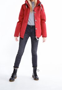 Rød vandtæt jakke med hætte, sølv lynlås og knapper. Sammen med grå skjorte, mørke jeans og sorte snørebåndsstøvler.