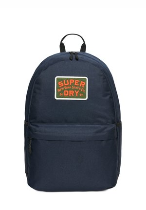 Granatowy plecak z plecioną teksturą, z przednią naszywką, na której widnieje napis "SUPER DRY" w czerwonym i zielonym, z zamykaną kieszenią i podwójnymi ramiączkami.