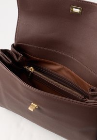 Smeđa teksturirana torba s prostranim unutrašnjim prostorom, koja sadrži zip džep i kopču od zlatnog hardvera. Glatka podstava upotpunjuje dizajn.