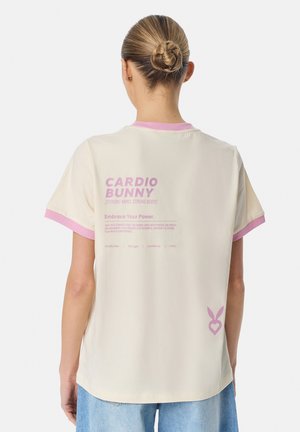 Naine, kellel on blondid juuksed kuklas, kannab kreemjat T-särki roosa äärise ja motiveeriva tekstiga, seisab seljaga vaataja poole ühtlasel taustal.