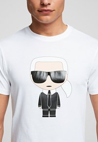 Camiseta blanca de algodón con un personaje de dibujos animados que lleva gafas de sol negras, cabello blanco y un traje negro, detallado con una corbata negra y cuello de camisa.