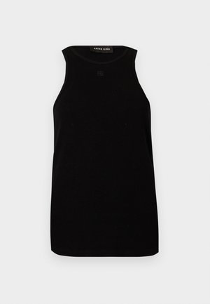 Femme debout portant un haut noir sans manches, un pantalon blanc à jambes larges avec une ceinture noire et des chaussures noires à bouts pointus sur fond blanc.