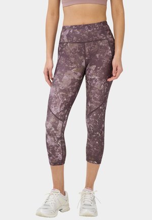 Vrouw die paarse gebloemde korte legging draagt en witte hardloopschoenen, staand tegen een effen achtergrond.