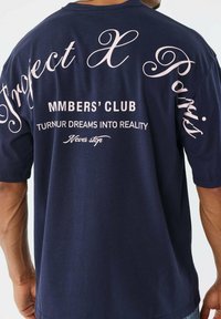 Man in een marineblauwe shirt met roze tekst en blokletters met de opschriften "Project X Paris," "MMBERS' CLUB," "TURNUR DREAMS INTO REALITY," en "Never stop."