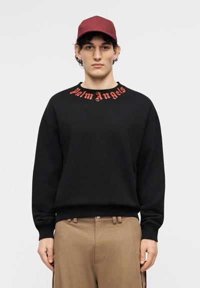 Νεαρός άνδρας που φοράει μωβ καπέλο, μαύρο φούτερ με κόκκινο κείμενο «Palm Angels» στο γιακά και καφέ παντελόνι, κοιτάζοντας μπροστά μπροστά σε ένα απλό φόντο.