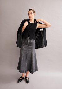 Gilet noir en tricot avec fermeture à bouton, associé à une longue jupe en denim grise. Des chaussures noires à bout pointu avec un détail de sangle complètent la tenue.