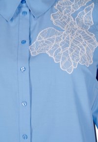 Camicia col colletto azzurro con ricami floreali bianchi in pizzo sul lato sinistro superiore. I bottoni sono azzurri e ben distanziati sulla parte frontale.