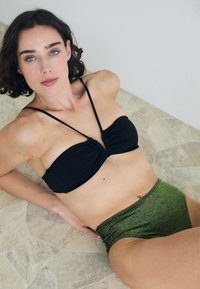 Juodas raukšlėtas bikini viršus su plonais petnešėlėmis, derinamas su aukštu liemeniu žaliais blizgučiais apatiniais, esant akmeniniu tekstūriniu paviršiumi.