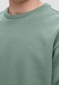 Sudadera verde de cuello redondo hecha de una tela suave, con un collar ribeteado y un sutil logo "QS" bordado en el mismo color.
