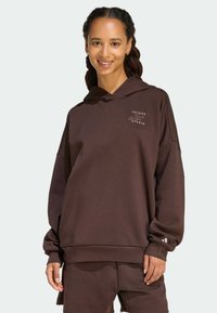 Donna che sorride, indossando un hoodie Adidas marrone scuro e pantaloni corti abbinati, in piedi contro uno sfondo chiaro e semplice.