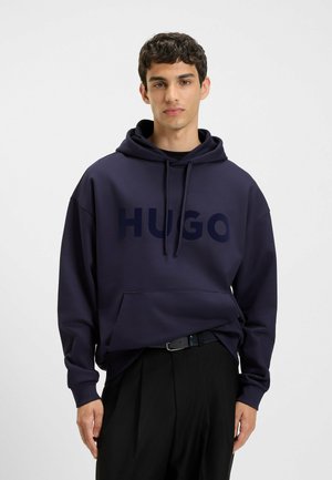 Young man wearing a navy blue hoodie with "HUGO" text, black pants, and a black belt, standing against a plain white background.
