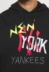 Černá mikina s kapucí, která má výrazný grafický design s nápisem "NEW YORK" v bílé a červené barvě a "YANKEES" v bílé, doplněná jasně žlutými prvky.
