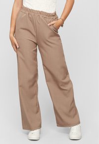 Beige, wijde broek met een elastische tailleband en zijzakken, gemaakt van een gladde stof. Gepaard met witte sneakers.