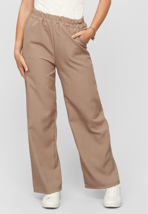 Pantalons beige à jambes larges avec une taille élastique et des poches latérales, fabriqués en tissu lisse. Associés à des baskets blanches.