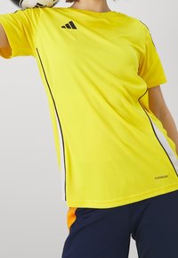 Žluté sportovní tričko s černými akcenty a logem. Vyrobeno z materiálu odvádějícího vlhkost, s kulatým výstřihem a krátkými rukávy.