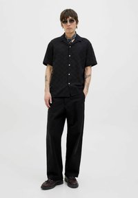 Schwarzes kurzärmliges Hemd mit Rautenmuster und Knöpfen, kombiniert mit lockeren schwarzen Hosen und dunklen Schuhen. Zubehör umfasst Sonnenbrille und ein Halstuch.