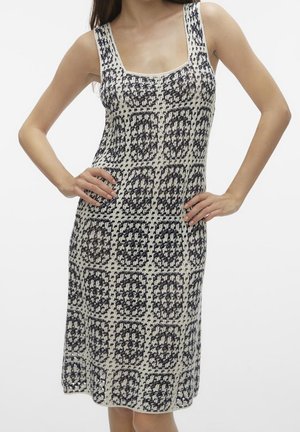 Kobieta w czarno-białej, bezrękawnikowej sukience z dzianiny na szydełku, o kwadratowym dekolcie, stojąca z rękami na biodrach na jednolitym tle.