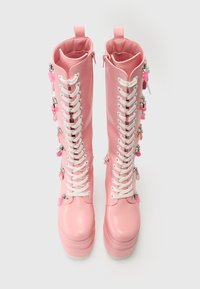 Bottes roses montantes en matériau brillant, dotées de lacets blancs, de semelles épaisses et de breloques décoratives sur les côtés.